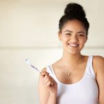 Brunette girl in a white tank top smiles while holding her toothbrush to care for her oral health