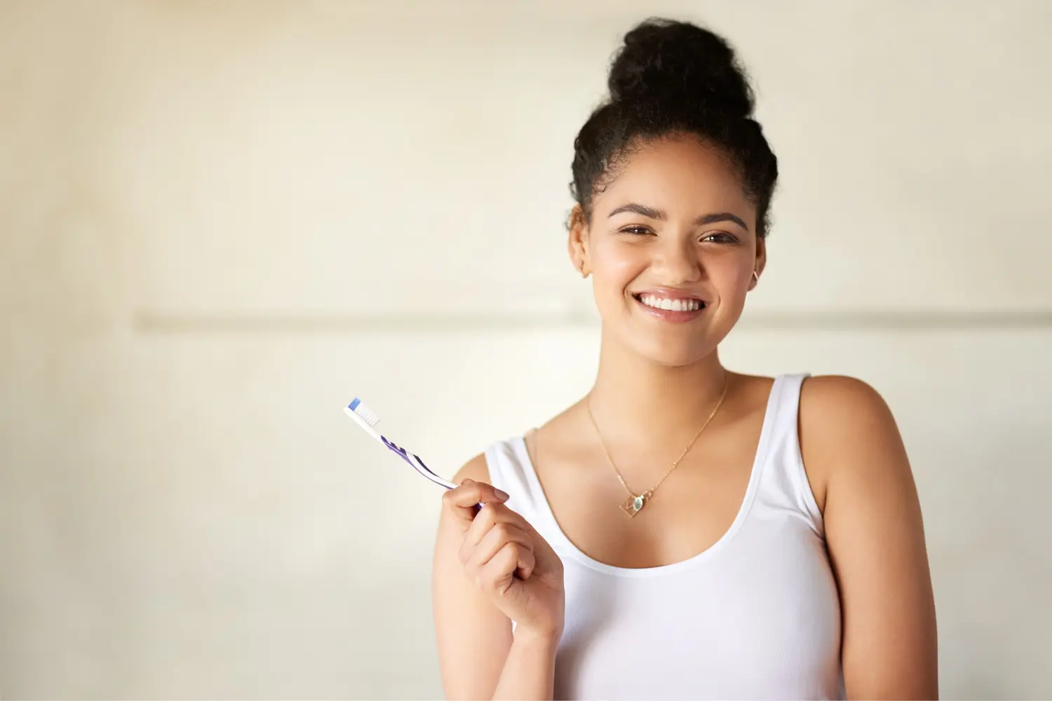 Brunette girl in a white tank top smiles while holding her toothbrush to care for her oral health