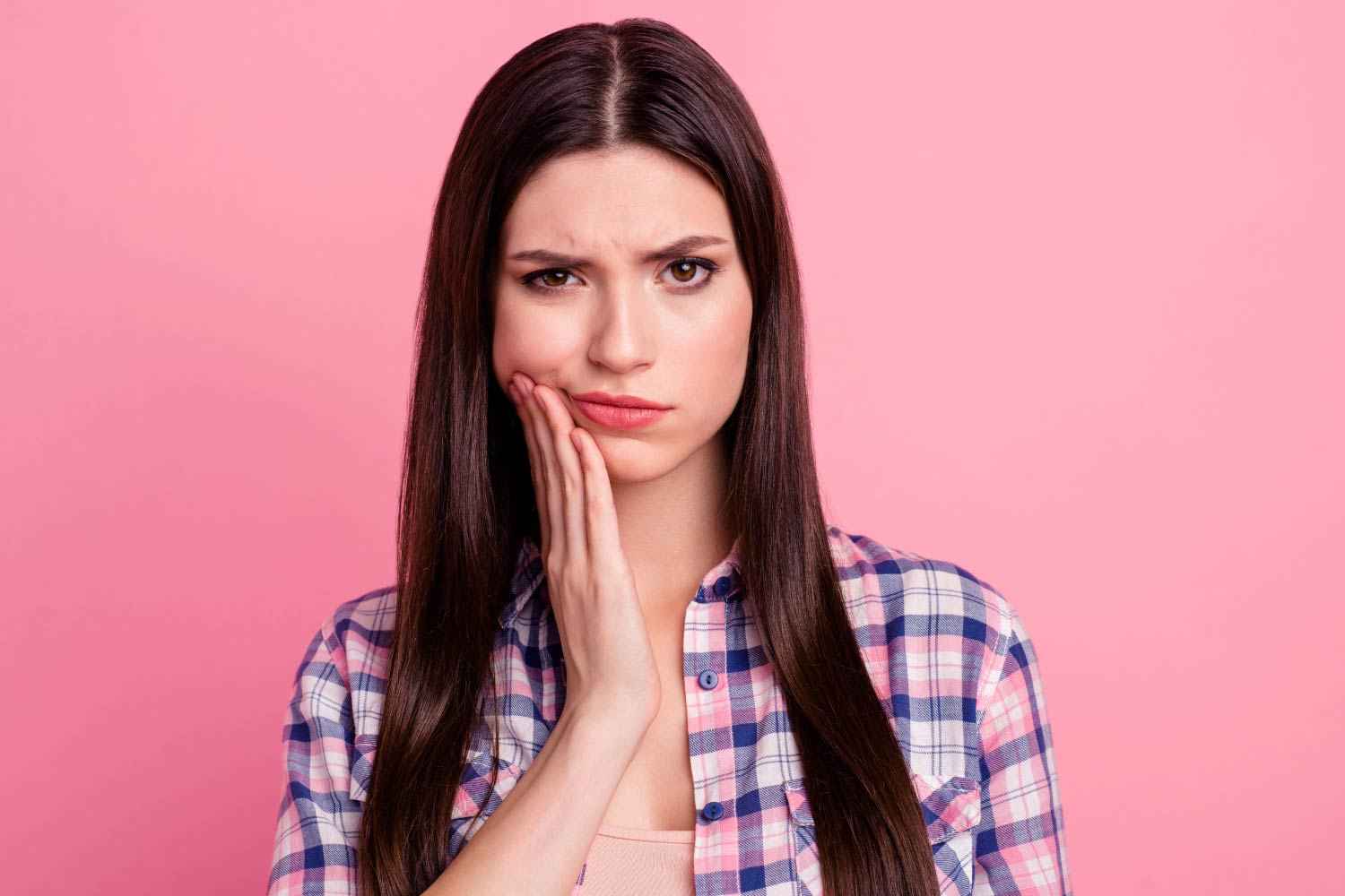 brunette woman with sensitive teeth cringes in pain and touches her cheek against a pink background