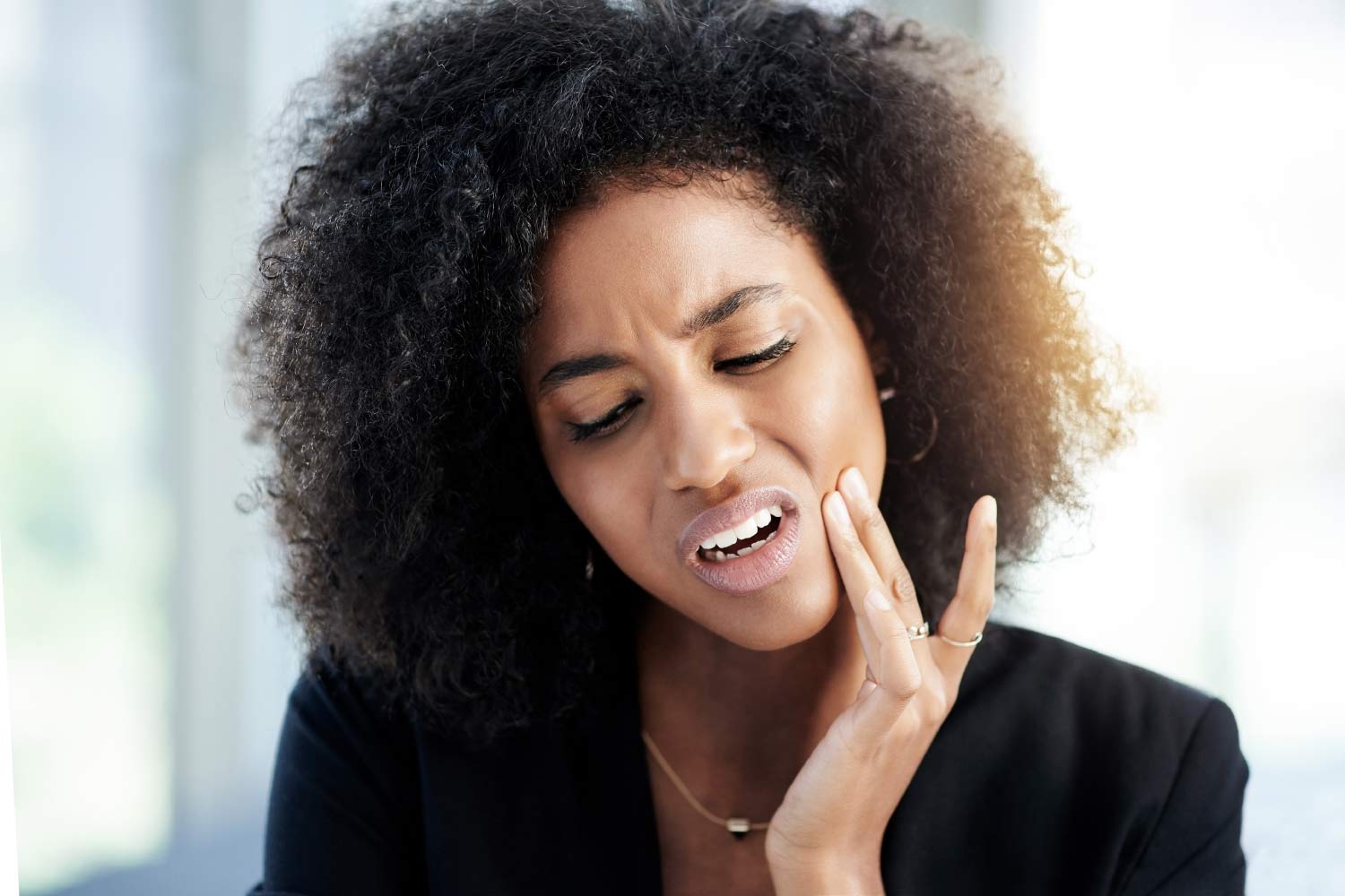 Curly-haired woman in a black shirt cringes in pain and touches her cheek due to a toothache dental emergency