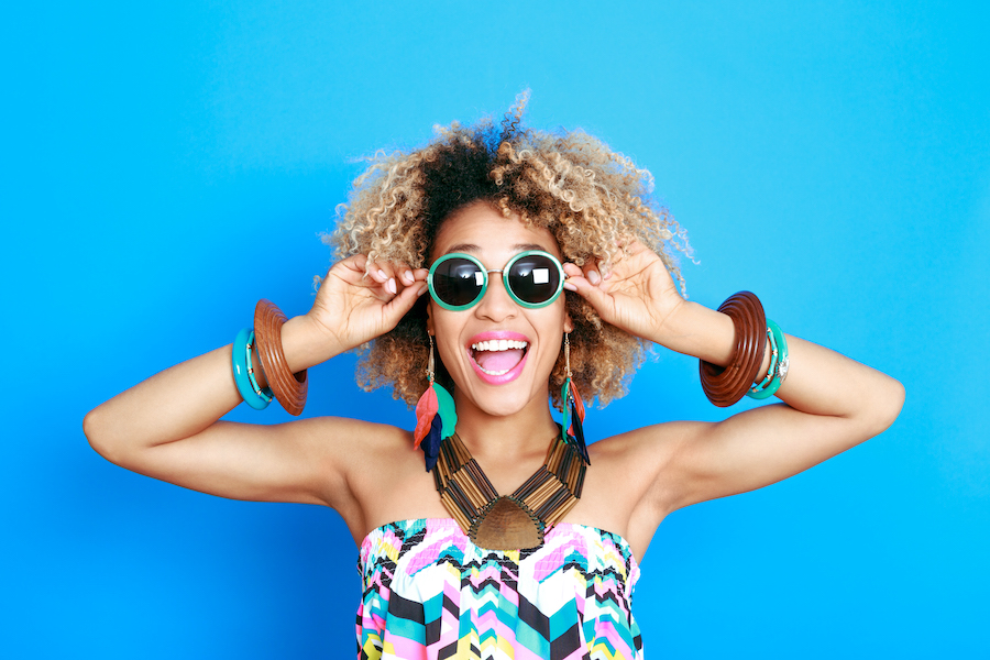 Black woman in summer clothes smiles while wearing sunglasses against a blue wall