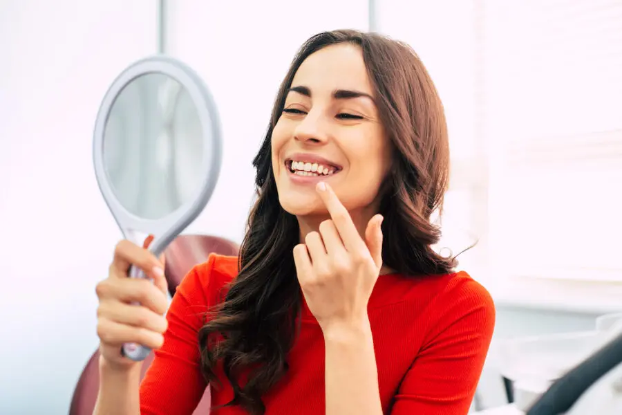 Brunette woman in a red blouse smiles at herself in a handheld mirror after teeth whitening