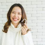 Asian woman in a white turtleneck sweater smiles as she holds up her Invisalign tray