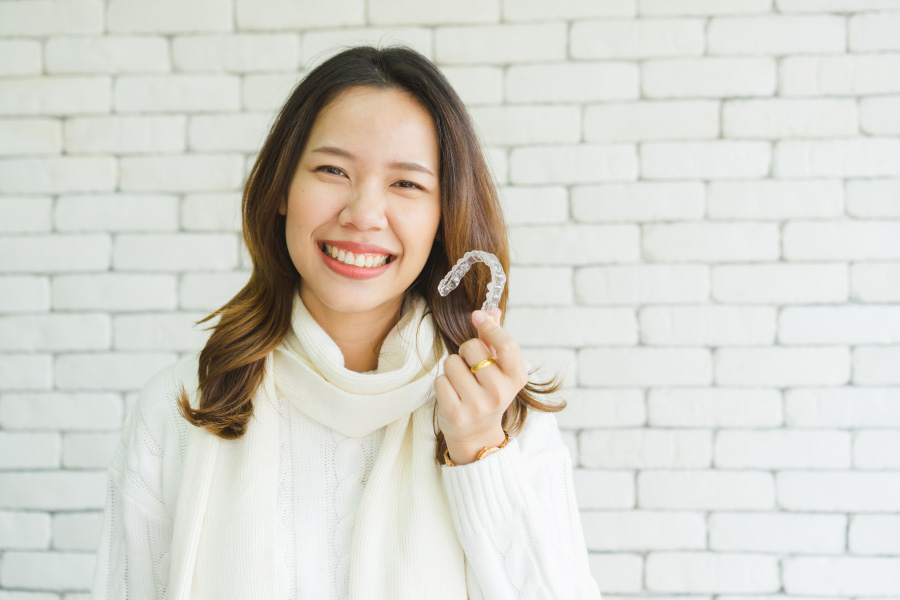 Asian woman in a white turtleneck sweater smiles as she holds up her Invisalign tray