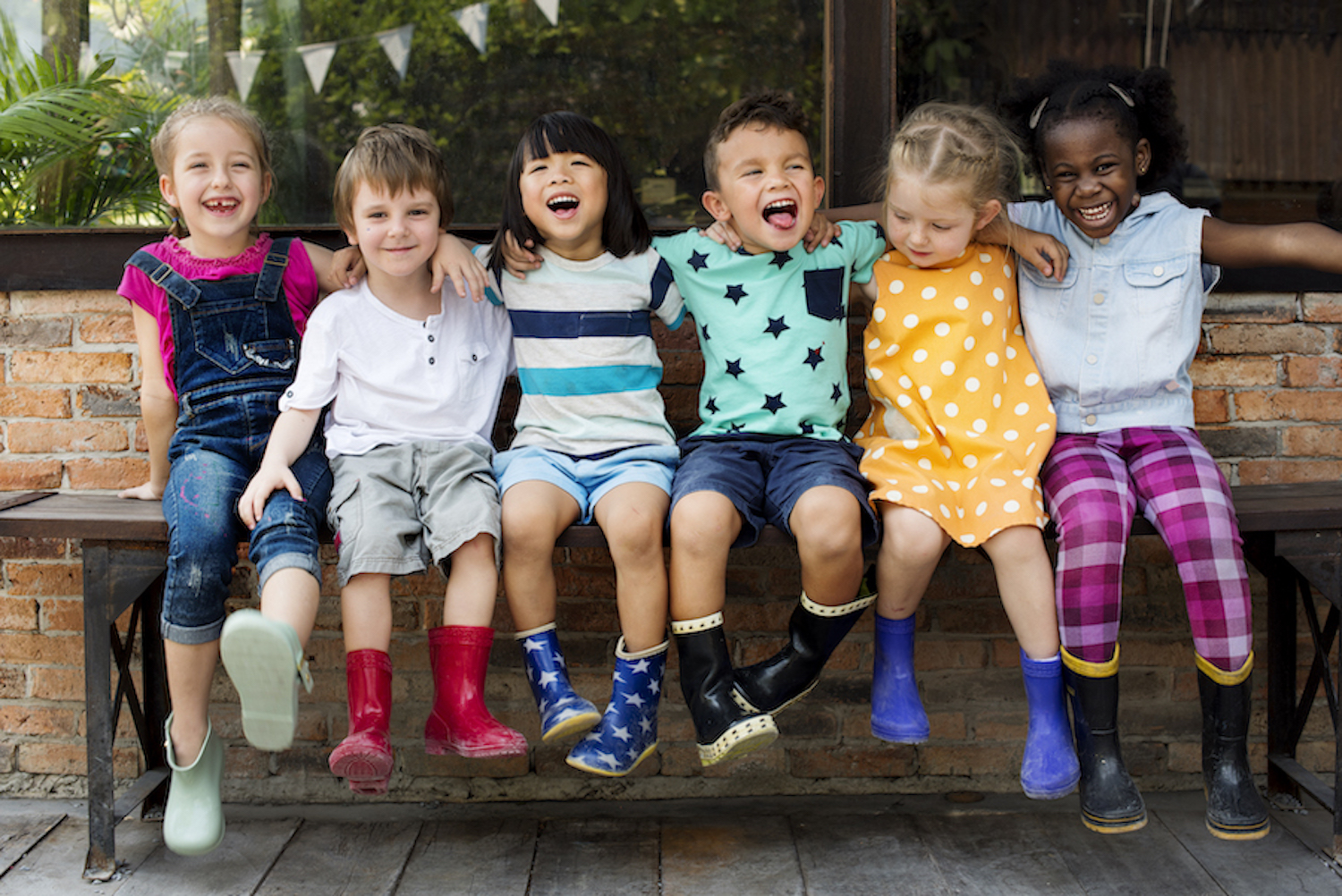 A group of young children sit on a bench together and smile with their arms around each other