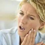 A blonde woman cringes in pain and holds her cheek due to a toothache
