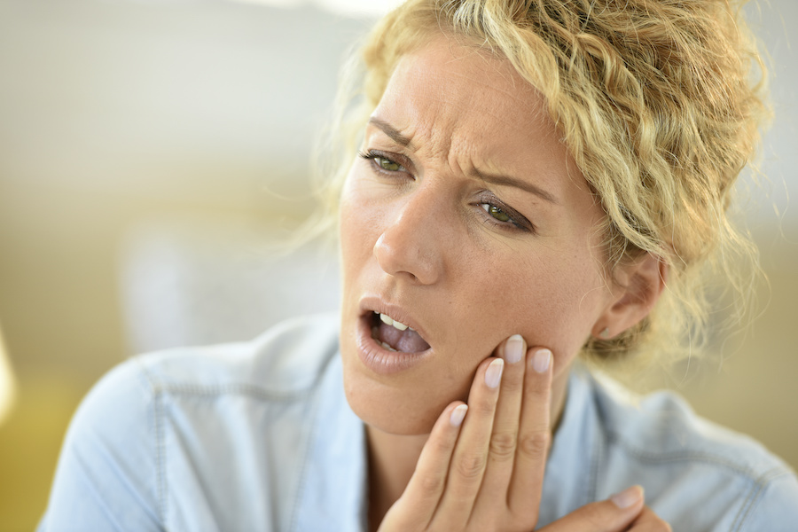A blonde woman cringes in pain and holds her cheek due to a toothache